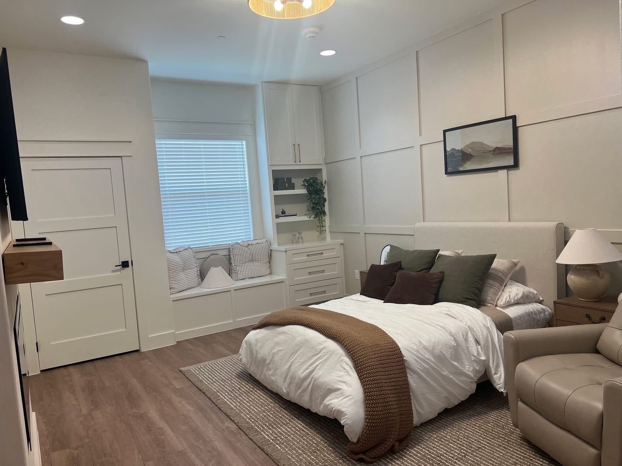 A calm resident bedroom with paneled walls, a window seat, built-in shelving, an upholstered bed, and a recliner.
